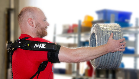 Effektiveres und gesünderes Arbeiten - mit dem Exoskelett MATE Mann in rotem T-Shirt trägt an den Schultern und Oberarmen ein schwarzes Exoskelett mit der Aufschrift MATE. Mit ausgestreckten Armen trägt er eine Schlauchrolle vor sich.
