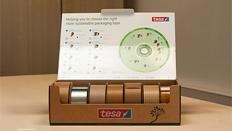 Pappkiste auf einem hellen Tisch, aufgeklappt. In der Kiste sieben Klebebandrollen, Infografik auf der Deckelinnenseite des Kartons.