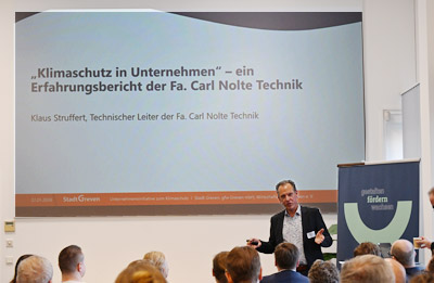 Auf einer Beamerleinwand ist ein Chart mit dem Titel "Klimaschutz im Unternehmen - ein Erfahrungsbericht der Fa. Carl Nolte Technik" zu sehen. Davor der Referent und einige Zuhörer, von denen man nur einen Teil der Köpfe sieht