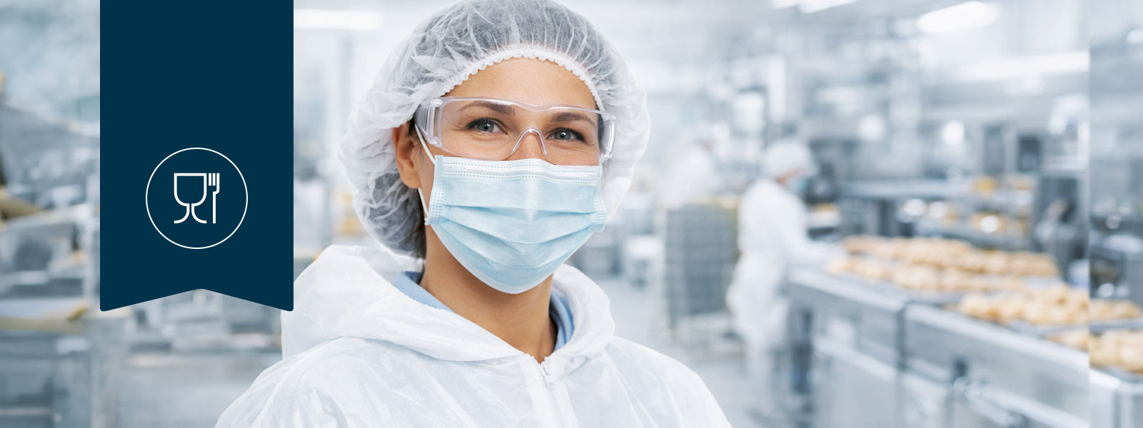 Junge Frau in einer Halle, in der Lebensmittel produziert werden. Sie trägt eine Hygienehaube, einen Mundschutz und eine Schutzbrille, schaut freundlich in die Kamera